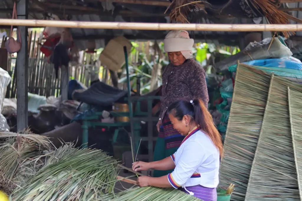 A Bulang woman smiling and weaving traditional straw mats, with her mother standing nearby watching.