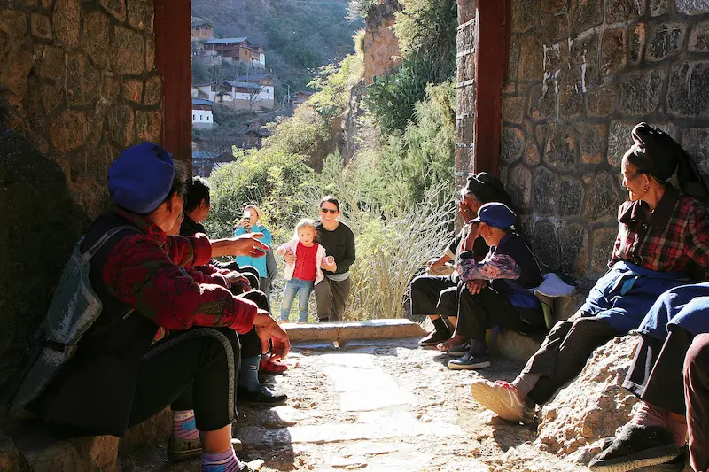 A German family walking through a stone gate while a group of local Naxi women sit nearby and watch.