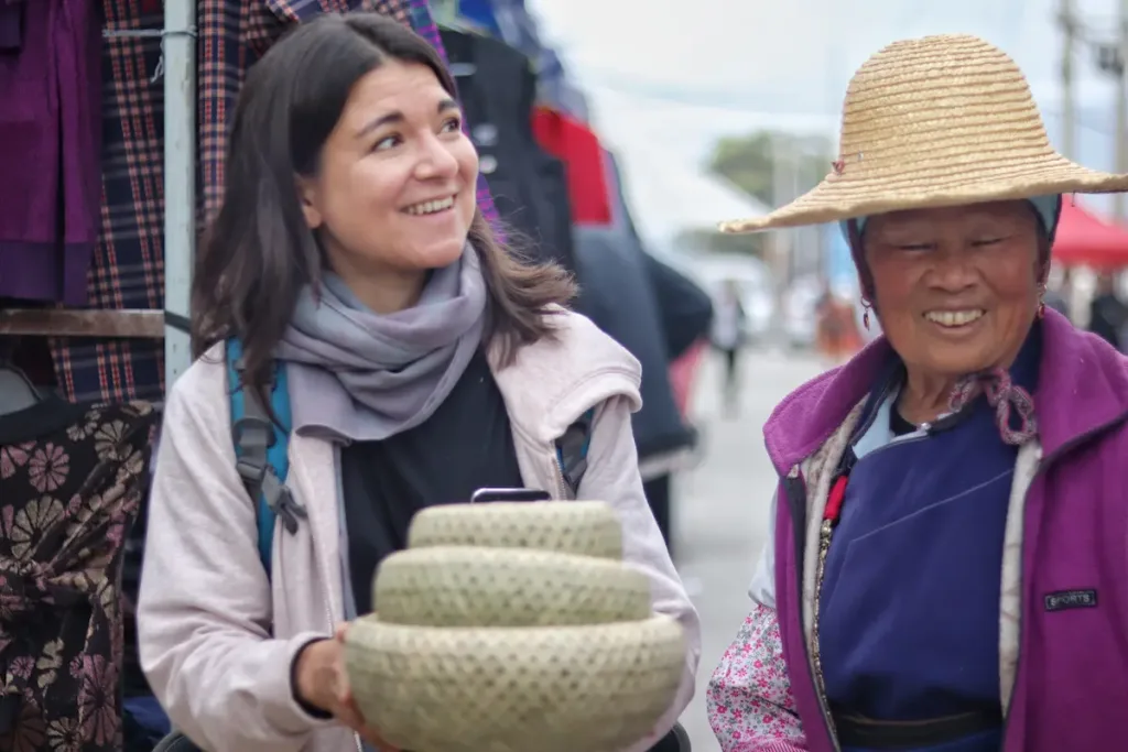 A German female guest holding bamboo bowls alongside a local woman at a market in Yunnan.