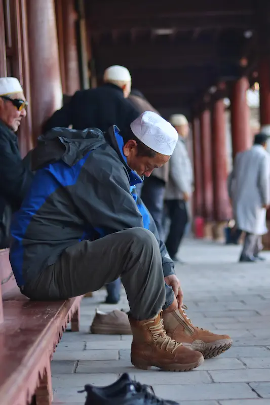 A Hui Muslim man wearing a white cap and sitting on the threshold of a prayer hall, putting on his shoes after prayer.