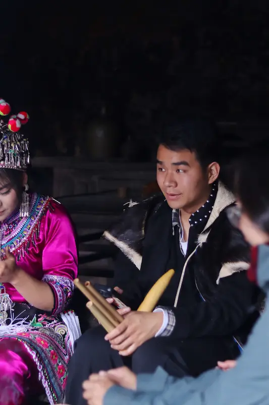 A Yi man holding a traditional musical instrument and looking into the distance, with a Yi girl in traditional clothing sitting beside him.