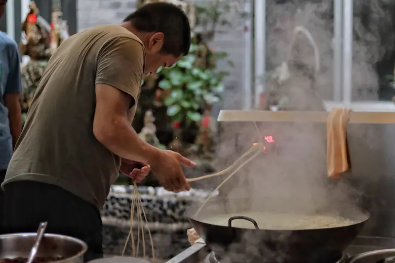 A man preparing a single strand of noodle in boiling water.