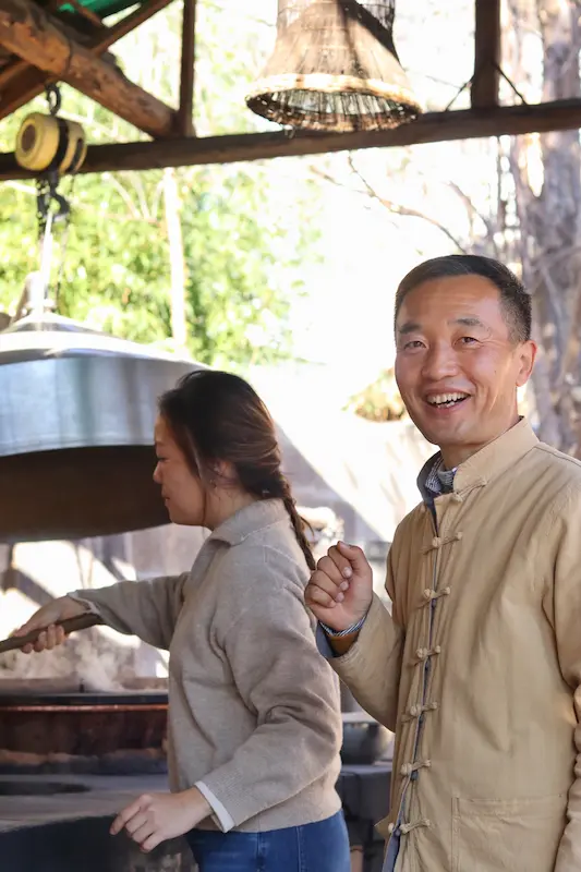 A pear artisan smiling with a Malaysian guest in the background, working on pear paste.