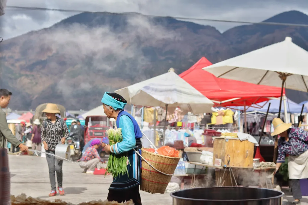 An elderly Bai minority woman walking through a local farmer's market in Yunnan, with mountains in the background.