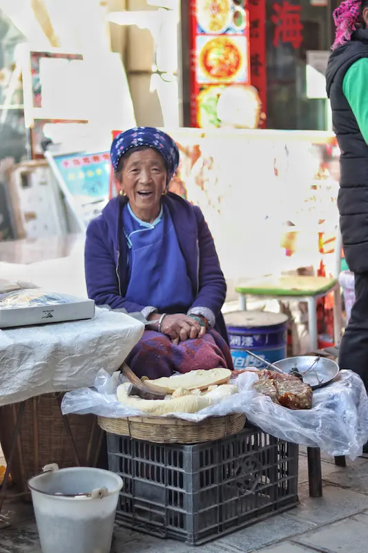 Bai ethnic woman smiling at her street food stall in a Yunnan market.
