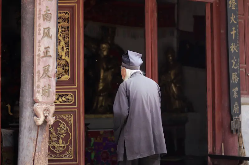 Elderly Taoist visitor in traditional robe and cap entering a temple hall at Weibaoshan, Weishan, Yunnan.
