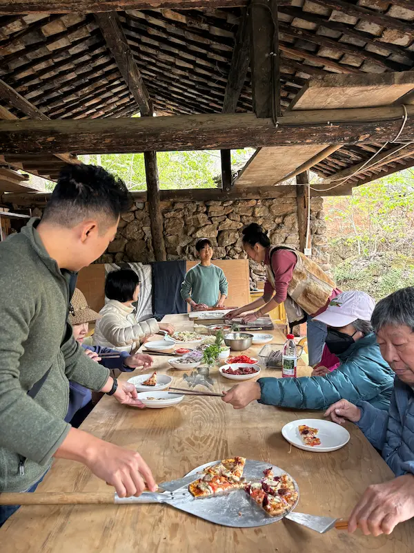 Group sharing freshly baked pizza at a long wooden table at a permaculture farm near Lijiang, Yunnan.
