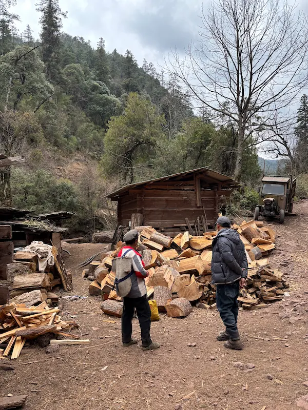 Lisu guide and a local villager standing near a wooden hut and stacked firewood in the mountains near Lijiang, Yunnan