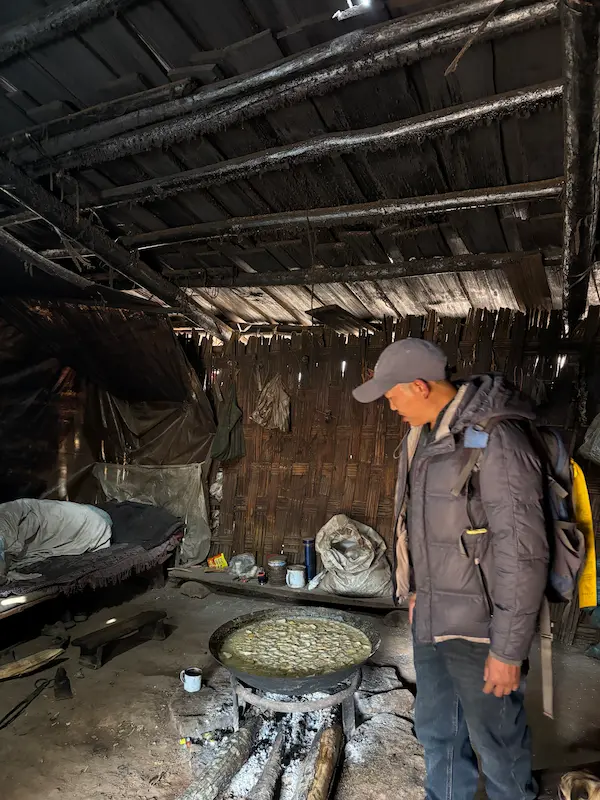 Lisu guide standing beside a traditional wood-fired stove inside a rustic forest hut near Lijiang, Yunnan