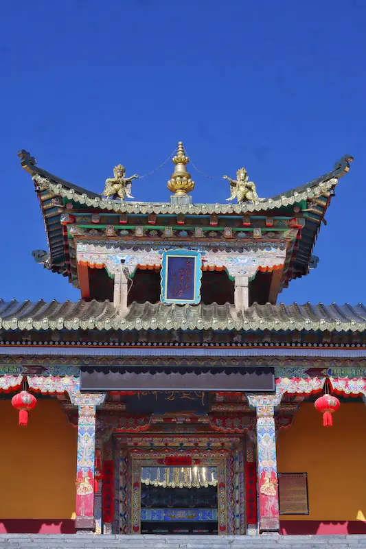 Traditional Chinese temple gate in Yunnan with ornate red and gold roof carvings, ceremonial guardian statues, and red lanterns against a blue sky.