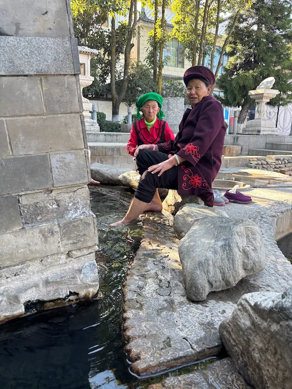 Two elderly Bai people looking at the camera while enjoying a foot soak in a natural hot spring in Dali.