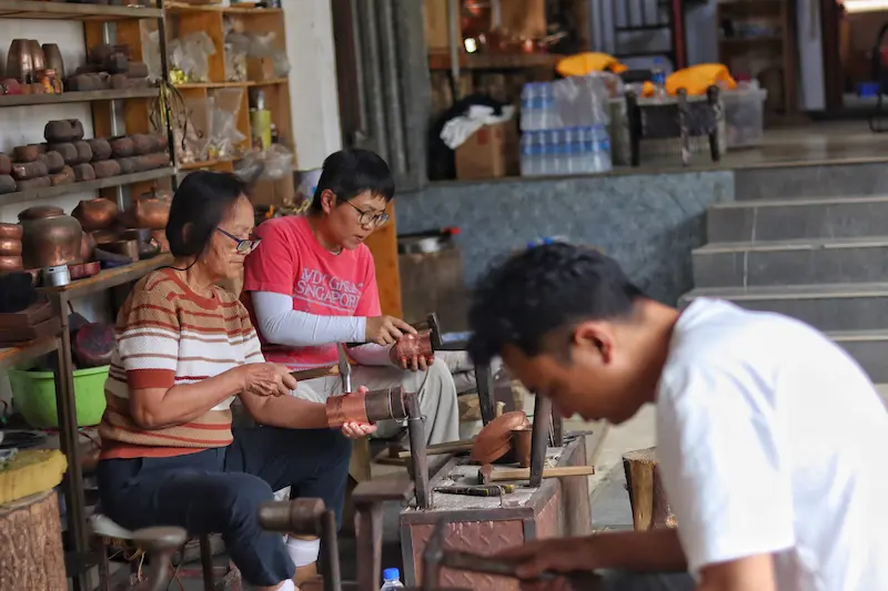 Two visitors learning traditional copper craftsmanship from a local artisan in Heqing, Dali, Yunnan.