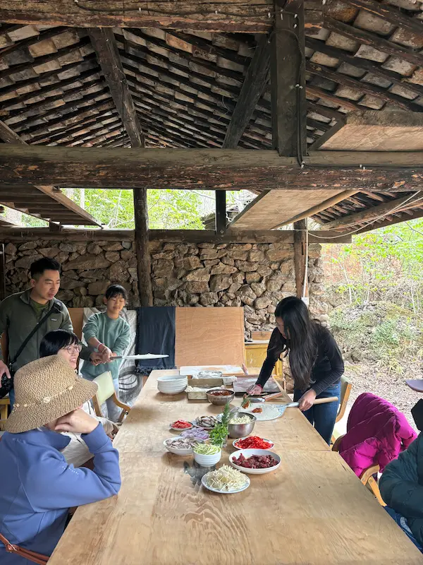 Visitors making homemade pizza with fresh toppings under a wooden barn structure at a permaculture farm near Lijiang, Yunnan.
