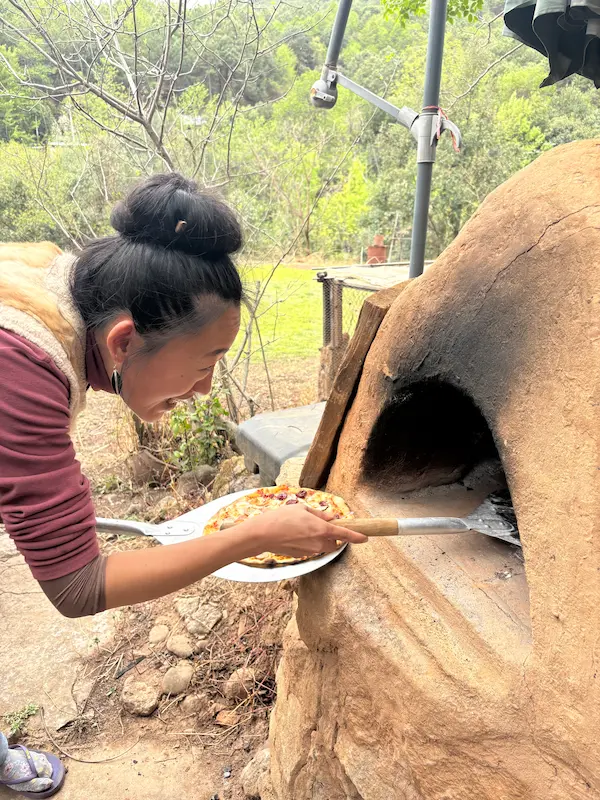 Woman placing a homemade pizza into a clay outdoor oven at a permaculture farm near Lijiang, Yunnan.