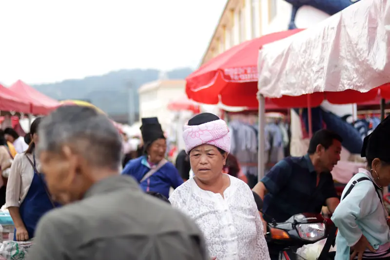 Busy Tengchong ethnic farmers' market with colorful umbrella stalls and a local Dai villager browsing the market