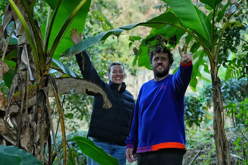 Two tourists waving and smiling in a lush Yunnan tea garden forest