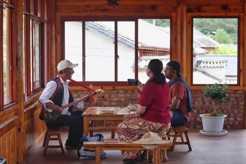 Two guests listening to a local musician play a traditional stringed instrument in a wooden house in Yunnan